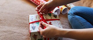 Eine Person sitzt auf dem Boden und zieht die rote Schleife eines Geschenkes fest. Im Hintergrund sind Geschenkpapierrollen. 