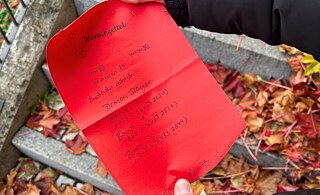 A red wishlist is held in one hand. Three book names are written on it. Autumn leaves can be seen in the background.