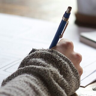 A person writing with a pen on paper, with a notebook and coffee cup in the background.