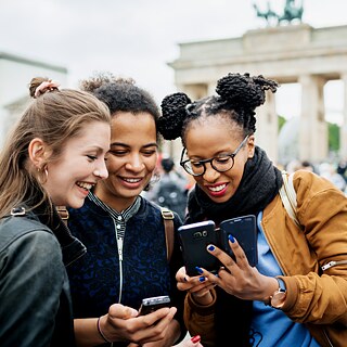 Drei junge Frauen stehen vor dem Brandenburger Tor in Berlin und betrachten lachend die Fotos auf einem Smartphone.