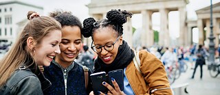 Drei junge Frauen stehen vor dem Brandenburger Tor in Berlin und betrachten lachend die Fotos auf einem Smartphone.