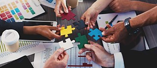  A group of people working together, each holding colorful puzzle pieces, symbolizing collaboration and teamwork in a professional setting with documents and charts on the table.