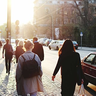 Menschen gehen bei Sonnenuntergang auf einem Bürgersteig in der Stadt, während Autos auf der Straße vorbeifahren.