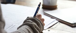 A person writing with a pen on paper, with a notebook and coffee cup in the background.