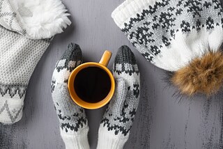 Fausthandschuhe halten eine heiße Tasse Tee und eine Wintermütze liegt daneben.