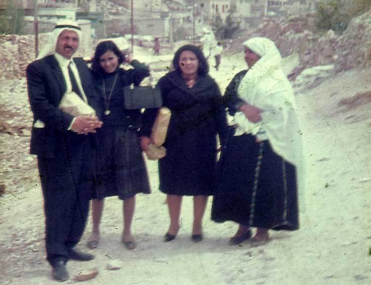 أربعة أشخاص على طريق صخرية في فلسطين العام ١٩٦٨. من اليسار إلى اليمين: رجل متوسط العمر يرتدي بزة رسمية وغطاء الرأس التقليدي بيده طرد، في المنتصف امرأتان بلباسهما، بيد إحداهما حقيبة، وبيد الأخرى بعض من الخبز. على اليمين، نساء يرتدين رداءً طويلاً أسود اللون وحجابًا طويلاً أبيض اللون.