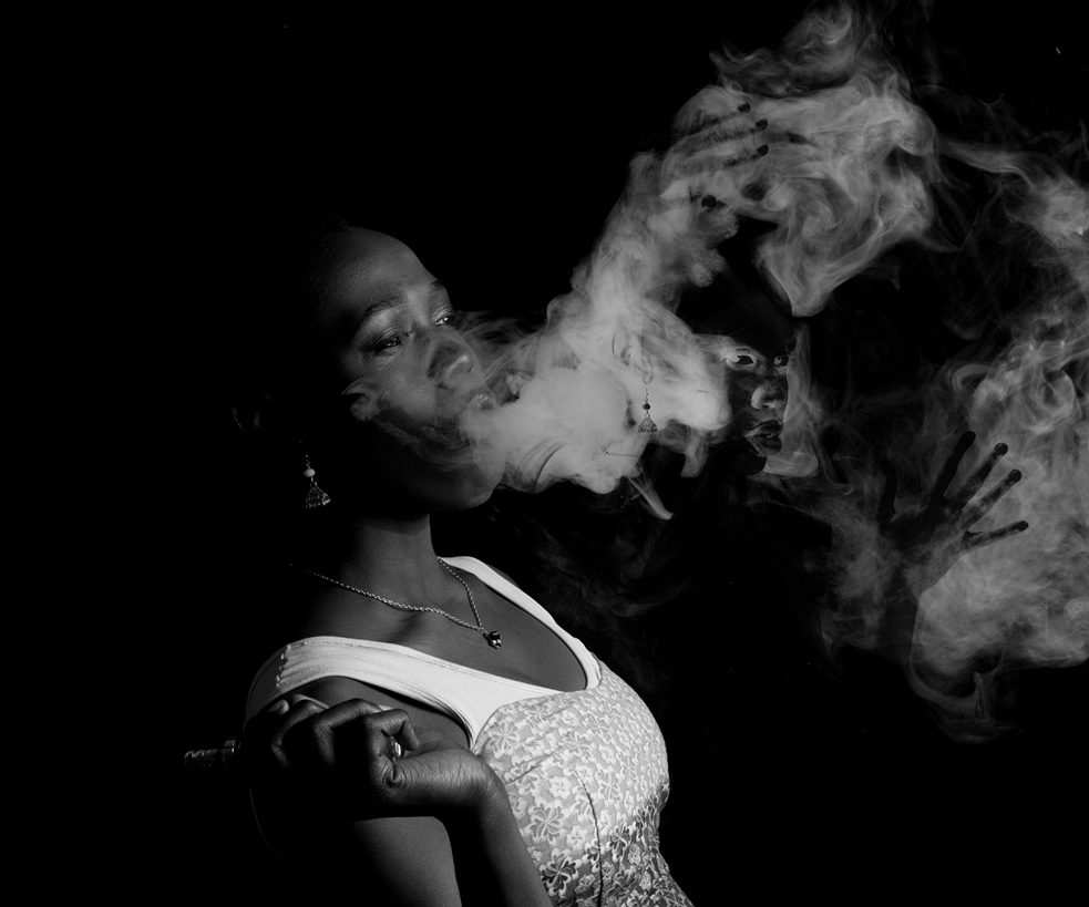 Photo en noir et blanc d'une femme regardant au loin, fumant, une image d'elle visible dans la fumée.