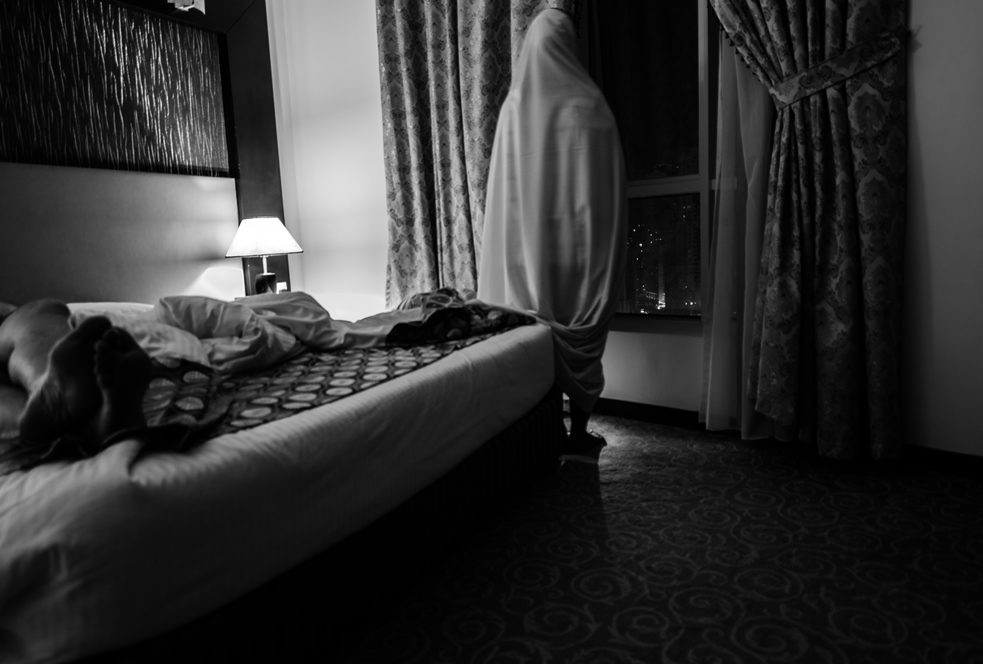 Photo en noir et blanc d'une pièce, une femme couverte debout près de la fenêtre regardant dehors, un homme allongé sur le lit.