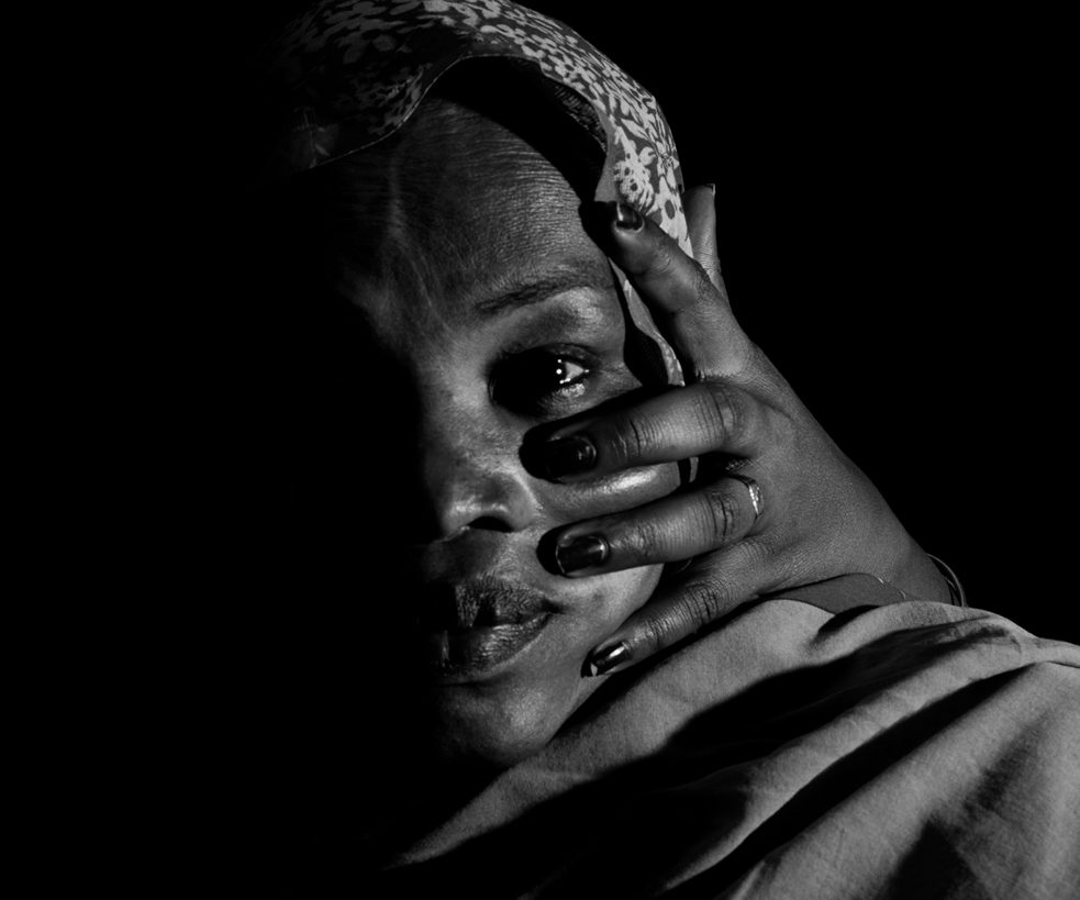 Photo en gros plan en noir et blanc du visage d'une femme, la moitié de son visage est dans l'ombre, de dos une main sur sa joue.