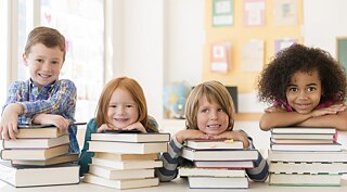 Vier Kinder sitzen in einem Klassenraum vor Bücherstapeln und lächeln. Sie stützen sich mit den Armen auf den Büchern ab.  © Goethe-Institut e.V.  Kinder lehnen auf Bücherstapeln