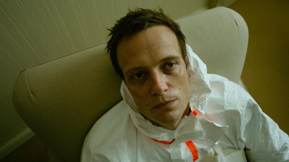 "Perfume - The Third Substance": close-up of Moritz de Vries (August Diehl) sitting on an armchair in a white protective suit, staring blankly into the camera.