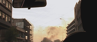 Durch eine Windschutzscheibe ist am Horizont Rauch zu sehen.  Am oberen Rand ist der Rückspiegel zu sehen. Das Foto wurde in einem urbanen Raum aufgenommen.