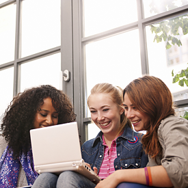 Drei junge Frauen sitzen in der Mediothek vor einem Laptop und lächeln.