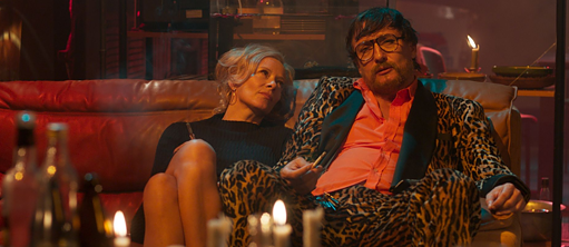 Un hombre con un traje de estampado leo y una mujer se sientan relajados en el sofá a la luz de las velas y fuman. 