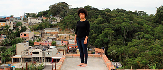 A foto mostra uma mulher negra de pé sobre um telhado de uma favela