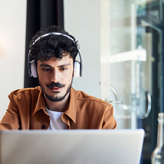 Un homme en situation d'apprentissage. Il est assis dans un café, porte des écouteurs et se concentre sur son ordinateur portable.