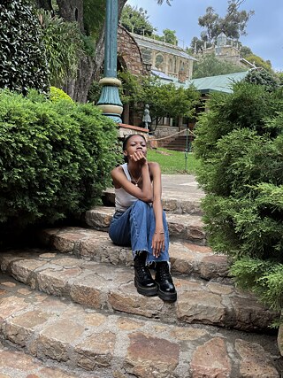 Moon Mokgoro sitting on natural stone steps in front of an old Art Nouveau house surrounded by bushes.