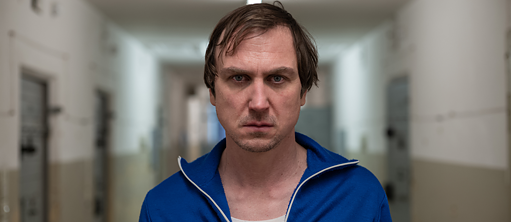 Close-up of a man wearing a blue tracksuit looking directly into the camera while standing in a prison gangway