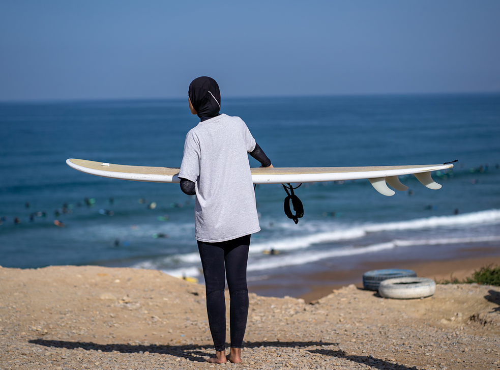امرأة شابة على الشاطئ مع لوح التزلج ووجهها إلى البحر