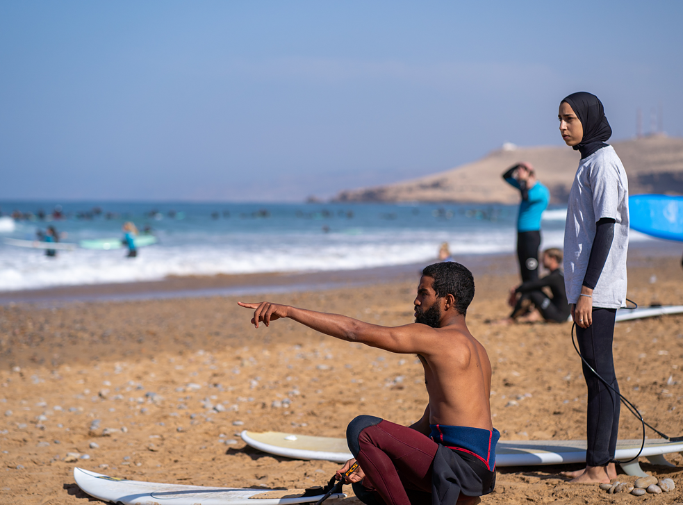 Redouane indique la direction de la mer et explique le comportement des vagues, Khadija écoute attentivement. Aujourd’hui, il surfe sur une shortboard, Khadi sur une longboard.