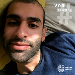 A portrait of a young man with short brown hair, five o’clock shadow and an earring can be seen lying on a bed. In the top right corner is a semi-transparent hashtag sign and above it the word Vorzeichen in white. In the bottom right corner is the logo of the Goethe-Institut. 