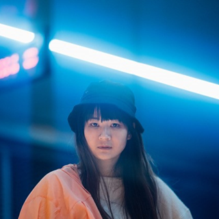 Die Künstlerin Edy Fung schaut direkt in die Kamera. Sie trägt einen schwarzen Hut und steht vor einem dunklen, verschwommenen Hintergrund. Im Hintergrund sind ein rotes Leuchtschild und eine bläulich-weiße Neonleuchte zu sehen. 