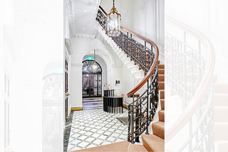 Brightly lit foyer depicting the large wrought iron door, two chandeliers, marble tile floor, a reception desk, and a grand stairway with a ornate iron and wood bannister. 