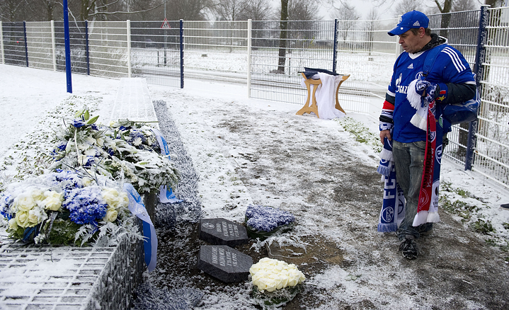 A fan of FC Schalke 04 stands at a grave in a jersey and draped with scarves and mourns 
