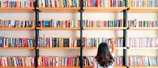 Eine Frau steht vor einem Bücherregal in einer Bibliothek und wählt ein Buch aus. Rückansicht.