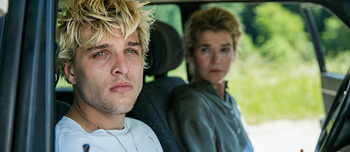 Ein junger Mann mit blonden Haaren und seine Mutter sitzen in einem Auto und schauen aus dem Fenster.
