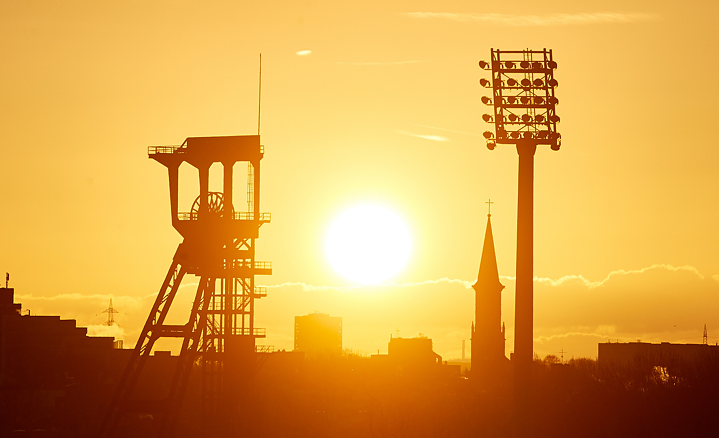 In the foreground a winding tower of the Holland colliery. In the background, a floodlight mast of the Lohrheide stadium at sunset.
