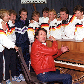 Die deutsche Fußballnationalmannschaft der Herren im steht in einem Musikstudio um ein Klavier, an dem der Schlagersänger Udo Jürgens sitzt.