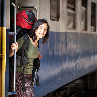 En ung kvinde med kort, brunt hår og en rød vandrerygsæk kigger ud af døren på et tog på stationen.