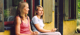 2 Frauen unterhalten sich auf einer Bank vor einem Haus, während eine raucht und die andere Kaffee trinkt