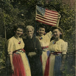 Das Bild zeigt eine Fotografie von vier Frauen, die historische Kleidung tragen, die die Farben der amerikanischen Flagge aufweist.