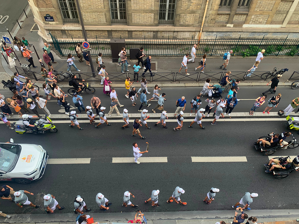 A street in Paris with a man carrying the Olympic flame. He is surrounded by many onlookers at the side of the road.