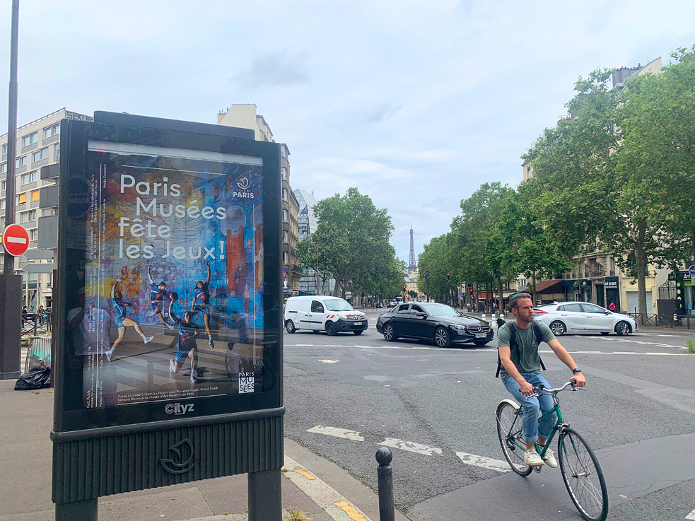 Ein Werbeplakat der Pariser Museen für ihr Sonderprogramm zu den Olympischen Spielen. Daneben ein Radfahrer, der um eine Kurve biegt. Im Hintergrund der Eiffelturm.