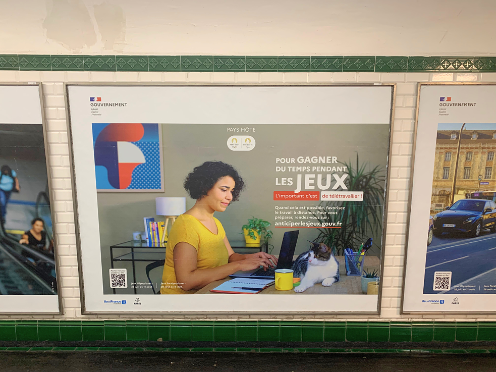 Ein Plakat in der Pariser Metro. Abgebildet ist eine Frau, die am Laptop arbeitet. Auf dem Schreibtisch sitzt eine Katze. Auf Französisch steht auf dem Plakat: "Um während der Spiele Zeit zu sparen, ist Telearbeit wichtig"