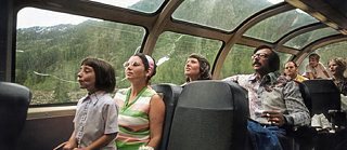 Auf dem Bild sieht man mehrere Menschen in einem Fahrzeug mit vielen Fenstern sitzen. Draußen sind Berge im Sommer zu sehen. Das Bild sieht retro aus.