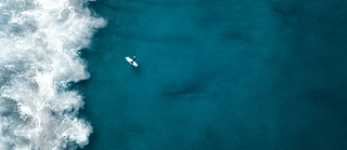 Auf dem Bild sieht man aus der Vogelperspektive ein türkisblaues Meer, einen Surfer und eine ankommende schäumende Welle.