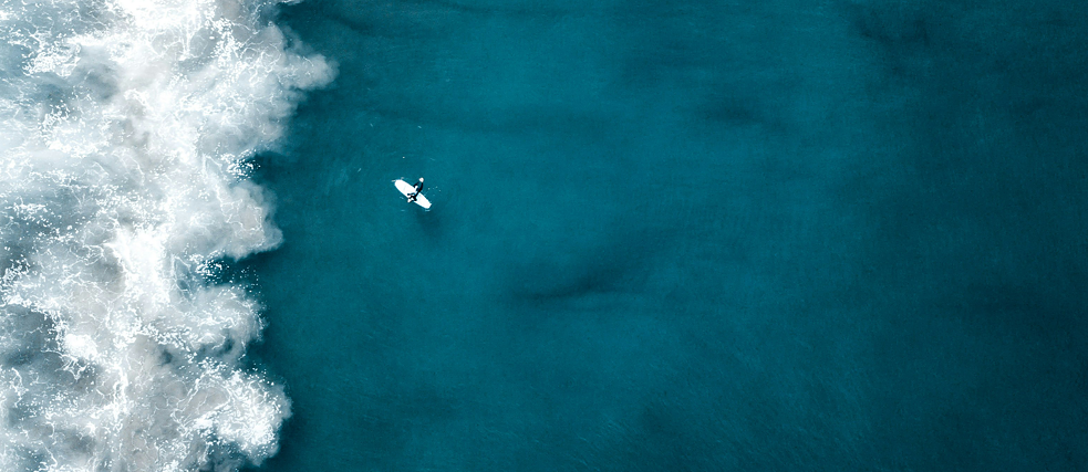 Auf dem Bild sieht man aus der Vogelperspektive ein türkisblaues Meer, einen Surfer und eine ankommende schäumende Welle.