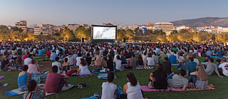 A large group of people sitting on a grassy field, watching an outdoor movie screening at dusk. © Goethe-Institut e.V. Eröffnung Infohaus & Filmvorführung