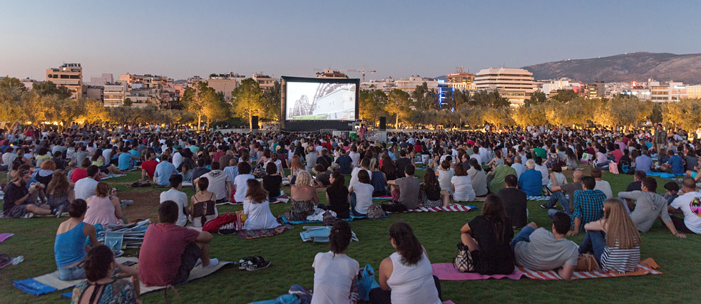 A large group of people sitting on a grassy field, watching an outdoor movie screening at dusk.