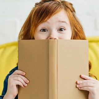 A young girl covers her face with a book, creating a playful and mysterious atmosphere in her surroundings.