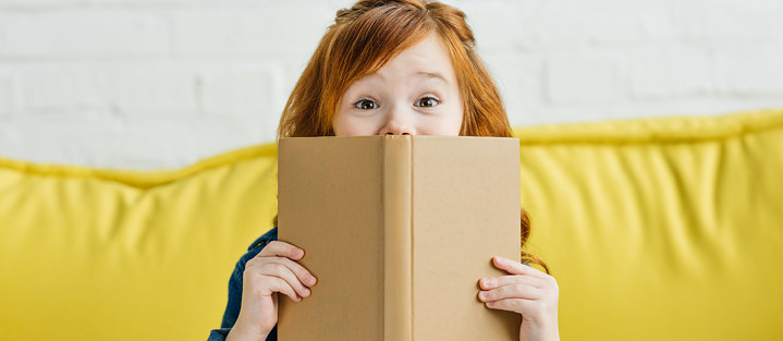 Ein junges Mädchen hält sich ein Buch vor das Gesicht und schaut gespannt in die Kamera