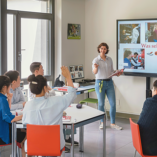 Eine Gruppe von Personen in einem Klassenzimmer vor einer Projektionsleinwand, die gemeinsam Deutsch lernen und diskutieren.