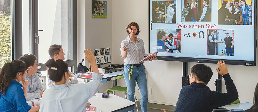 Eine Gruppe von Personen in einem Klassenzimmer vor einer Projektionsleinwand, die gemeinsam Deutsch lernen und diskutieren.