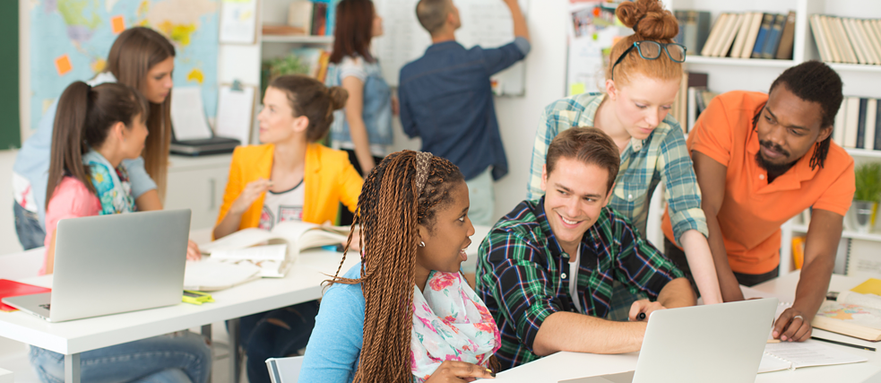  A group of diverse students collaborating in a busy classroom, with laptops, books, and a whiteboard, creating an active and engaging learning environment.