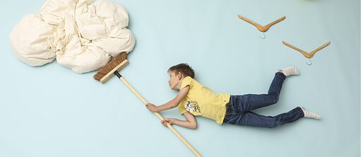 Foto von einem Kind mit gelben Tee-shirt, das auf einem helleblauen Fotohintergrund horizontal auf der Seite liegt und macht als ob er eine Wolke (dargestellt mit einer Daunendecke) mit einem Besen schieben würde. Zwei Bügel oben rechts stellen Vögel dar.  