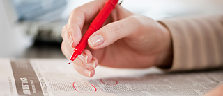 Close-up of a female hand marking areas in a newspaper with a red pen.